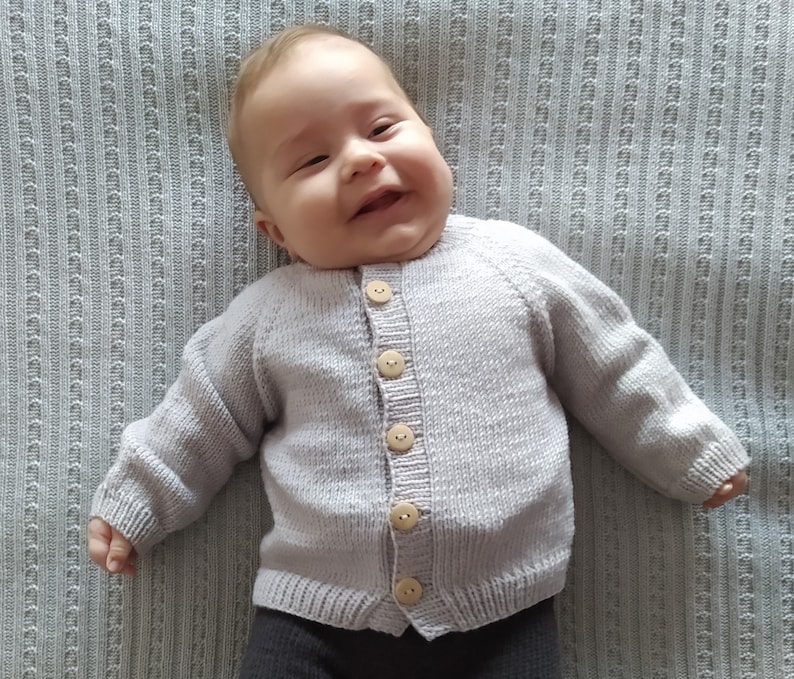 May include: A baby wearing a light purple knitted cardigan with wooden buttons. The cardigan has a ribbed texture and a classic button-up design.
