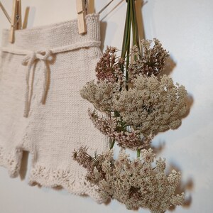 May include: A pair of white knitted shorts with a lace trim and a tie at the waist, hanging on a clothesline with dried flowers.