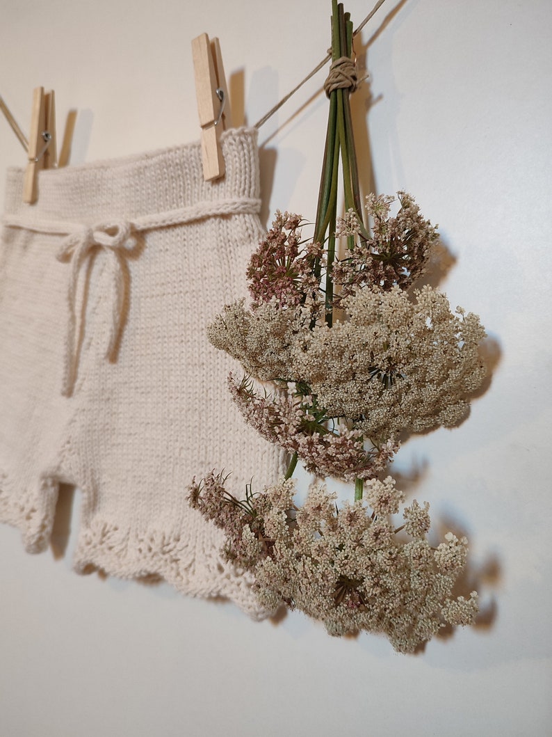 May include: A pair of white knitted shorts with a lace trim and a tie at the waist, hanging on a clothesline with dried flowers.