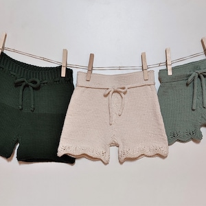 May include: Three pairs of knitted shorts hanging on a clothesline. The shorts are in green, beige, and a lighter shade of green. The shorts have a drawstring waist and a scalloped hem.
