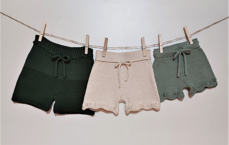 May include: Three pairs of knitted shorts hanging on a clothesline. The shorts are in green, beige, and a lighter shade of green. The shorts have a drawstring waist and a scalloped hem.
