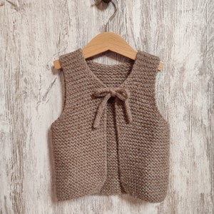 May include: A brown knitted vest with a bow tie detail hanging on a wooden hanger against a white wood background.