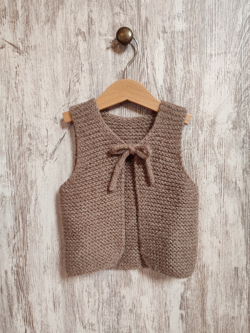 May include: A brown knitted vest with a bow tie detail hanging on a wooden hanger against a white wood background.