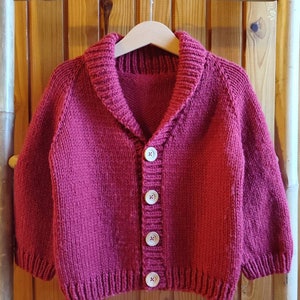 May include: A deep magenta knitted cardigan featuring a shawl collar, long sleeves with ribbed cuffs, and a front button closure with four light-coloured circular buttons. The garment is displayed on a wooden hanger.