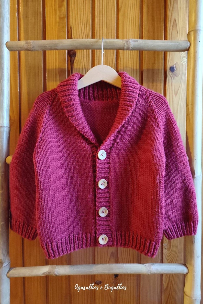 May include: A deep magenta knitted cardigan featuring a shawl collar, long sleeves with ribbed cuffs, and a front button closure with four light-coloured circular buttons. The garment is displayed on a wooden hanger.