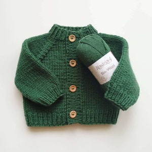 Puede incluir: Un c&aacute;rdigan tejido de color verde oscuro con mangas largas y cuello, pu&ntilde;os y dobladillo acanalados. El frente presenta cuatro botones de madera de color marr&oacute;n claro. Un ovillo de lana Ros&aacute;rios 4 Bio-Wool a juego descansa sobre la prenda.