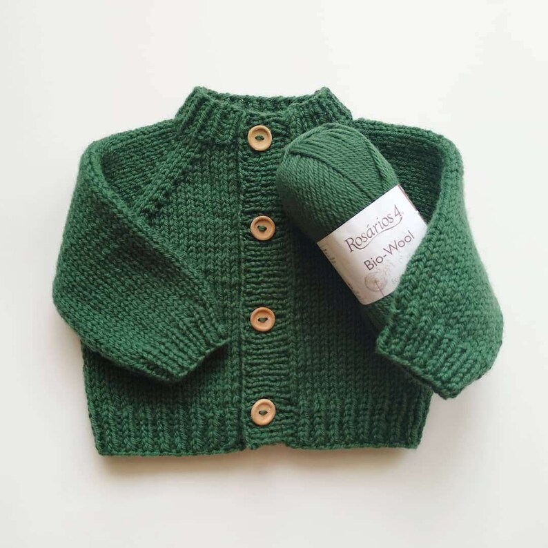 Puede incluir: Un c&aacute;rdigan tejido de color verde oscuro con mangas largas y cuello, pu&ntilde;os y dobladillo acanalados. El frente presenta cuatro botones de madera de color marr&oacute;n claro. Un ovillo de lana Ros&aacute;rios 4 Bio-Wool a juego descansa sobre la prenda.