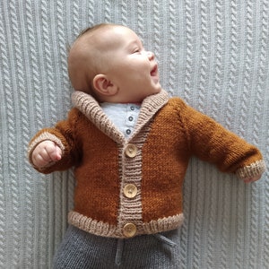 May include: A hand-knitted baby cardigan in a rich rust-brown colour featuring a contrasting beige shawl collar, cuffs, and hem. The garment is secured with three large, light-coloured wooden buttons down the front centre.