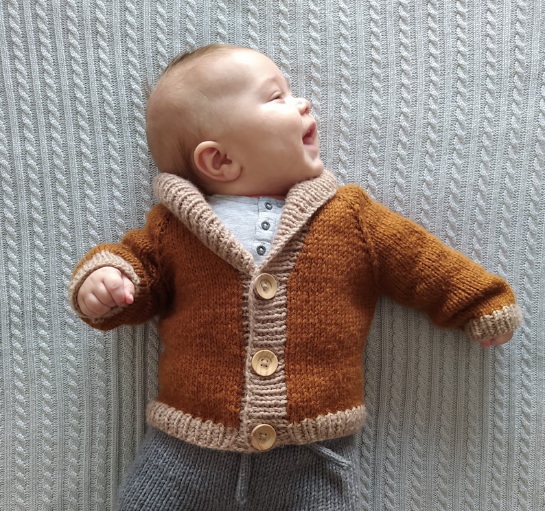 May include: A hand-knitted baby cardigan in a rich rust-brown colour featuring a contrasting beige shawl collar, cuffs, and hem. The garment is secured with three large, light-coloured wooden buttons down the front centre.