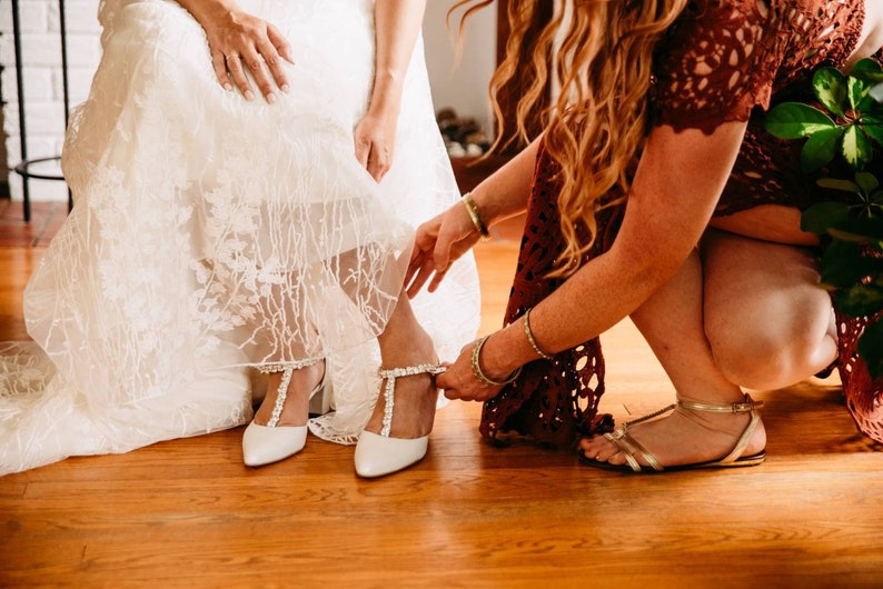 May include: A bride is having her white wedding shoes put on by a bridesmaid. The shoes have a pearl embellishment and a pointed toe.