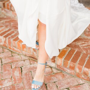 May include: A woman's foot wearing a light blue suede heeled sandal with a thin ankle strap. The sandal is on a brick walkway.