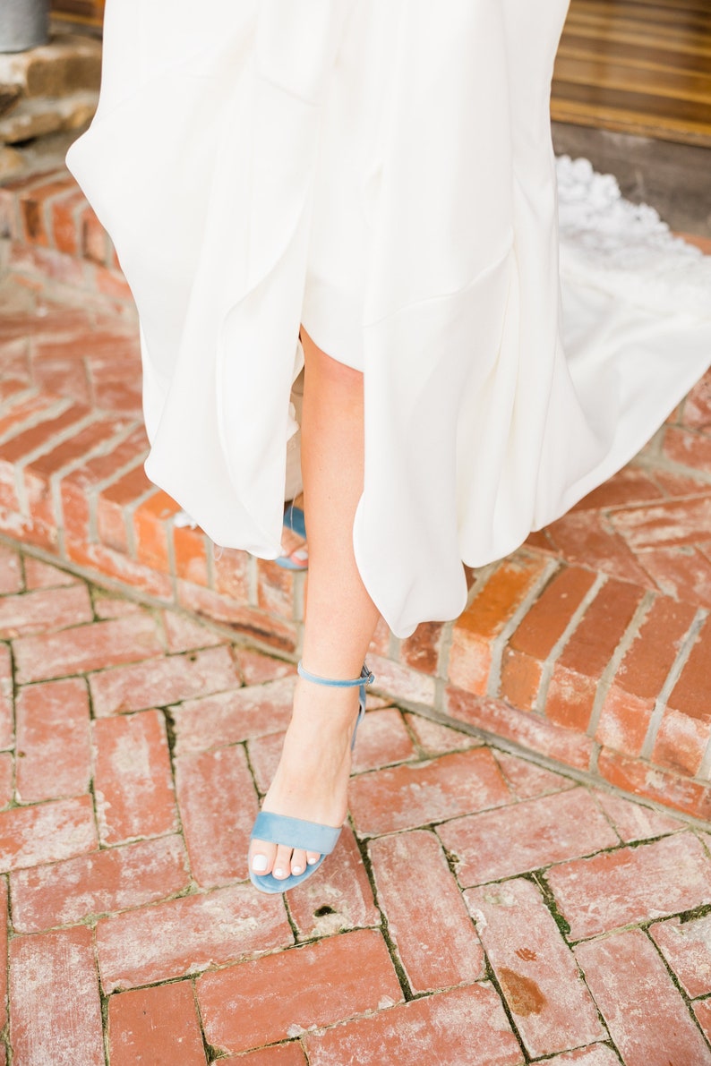 May include: A woman's foot wearing a light blue suede heeled sandal with a thin ankle strap. The sandal is on a brick walkway.