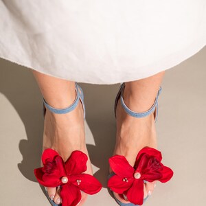A pair of light blue denim women's sandals adorned with vibrant red flower accents. Each flower features a pearl-like center detail, adding an elegant touch. The sandals have ankle straps and open toes, showcasing a summery style. These stylish shoes are perfect for adding a pop of color to any outfit.