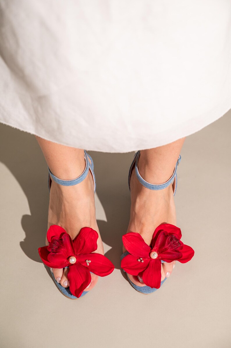 A pair of light blue denim women's sandals adorned with vibrant red flower accents. Each flower features a pearl-like center detail, adding an elegant touch. The sandals have ankle straps and open toes, showcasing a summery style. These stylish shoes are perfect for adding a pop of color to any outfit.