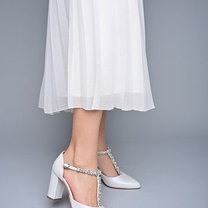 May include: White bridal shoes with a pointed toe and a chunky heel. The shoes have a T-strap with a crystal embellishment.