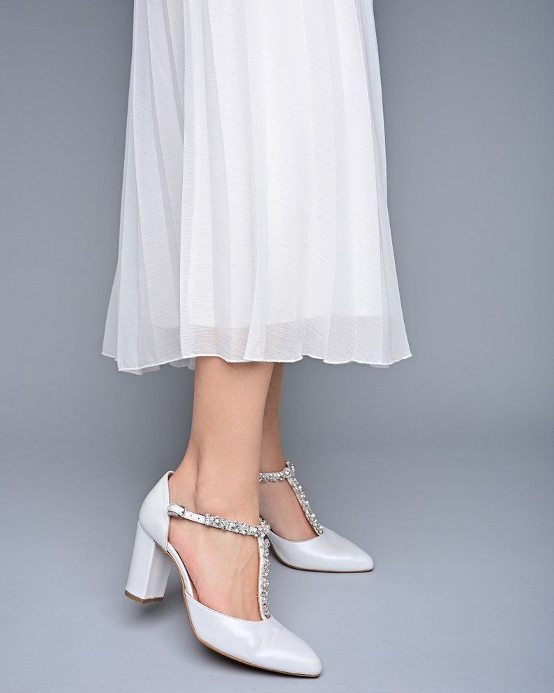 May include: White bridal shoes with a pointed toe and a chunky heel. The shoes have a T-strap with a crystal embellishment.