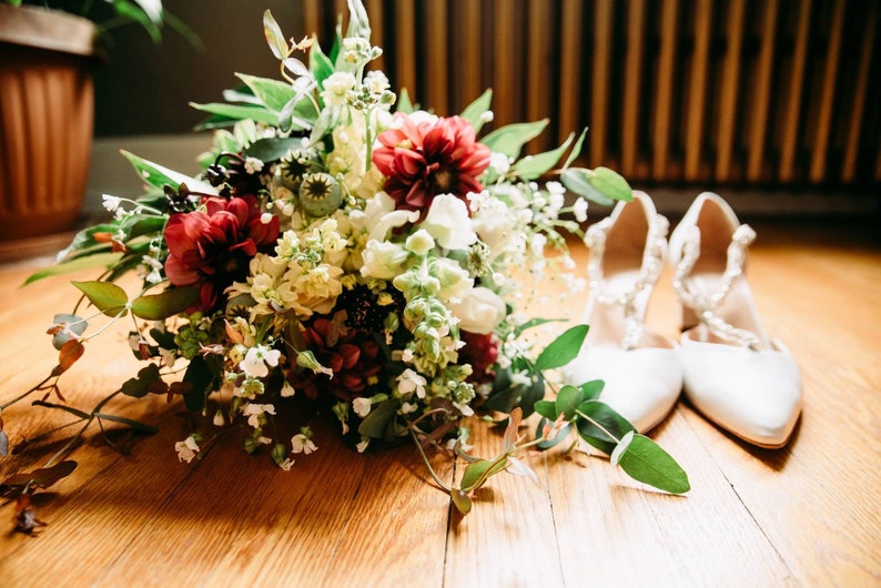 May include: A bridal bouquet with white, red, and green flowers, and a pair of white satin shoes.