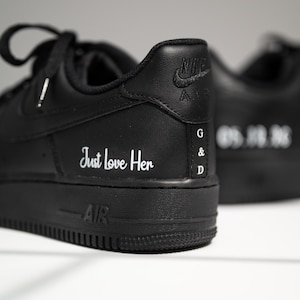 Könnte beinhalten: Schwarze Nike Air Force 1 Sneaker mit weißem Text "Just love Her" an der Seite. Die Ferse zeigt das Nike-Logo und die Initialen "G & D". Der andere Schuh ist leicht unscharf.
