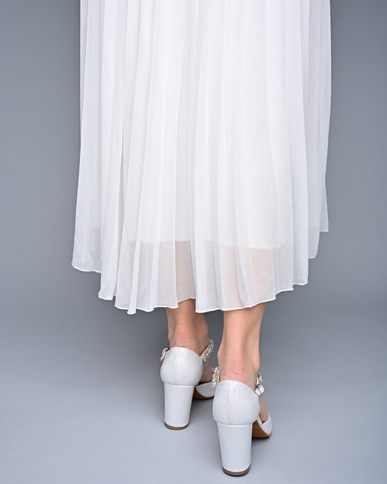 May include: White pleated skirt with white heeled shoes featuring a jeweled buckle.