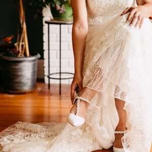 May include: A bride in a white lace wedding dress puts on a white bridal shoe with a pearl strap. The shoe has a pointed toe and a low heel.