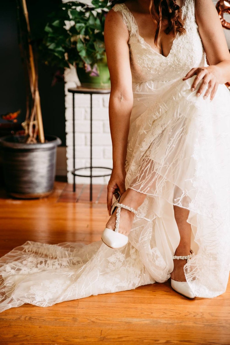 May include: A bride in a white lace wedding dress puts on a white bridal shoe with a pearl strap. The shoe has a pointed toe and a low heel.