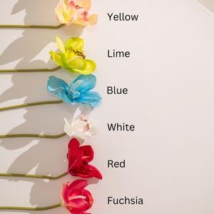 A row of artificial orchid stems in various colors, including magenta, yellow, lime, blue, white, red, fuchsia, and orange. Each stem is labeled with its corresponding color name. The image showcases a variety of vibrant hues and the artificial flowers' realistic appearance.
