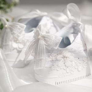 May include: White sneakers adorned with delicate lace, floral appliqués, and pearl accents. The shoes feature large, sheer bows and are designed with an elegant, bridal aesthetic. The word "AIR" is visible on the side.