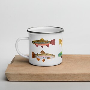 May include: A white enamel mug with a silver rim and a handle on the left side. The mug features a colorful illustration of several trout fish with detailed spots and fins in shades of orange, yellow, green, and brown against a white background.