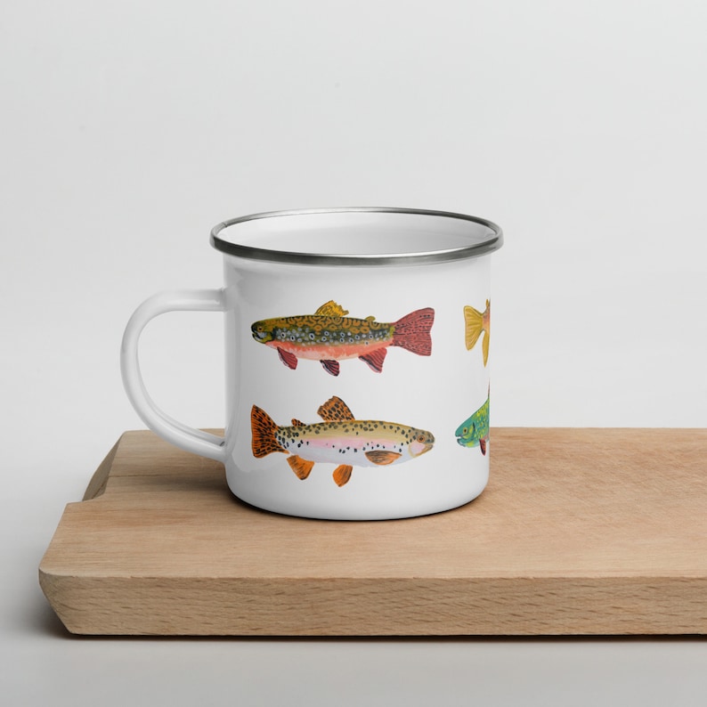 May include: A white enamel mug with a silver rim and a handle on the left side. The mug features a colorful illustration of several trout fish with detailed spots and fins in shades of orange, yellow, green, and brown against a white background.