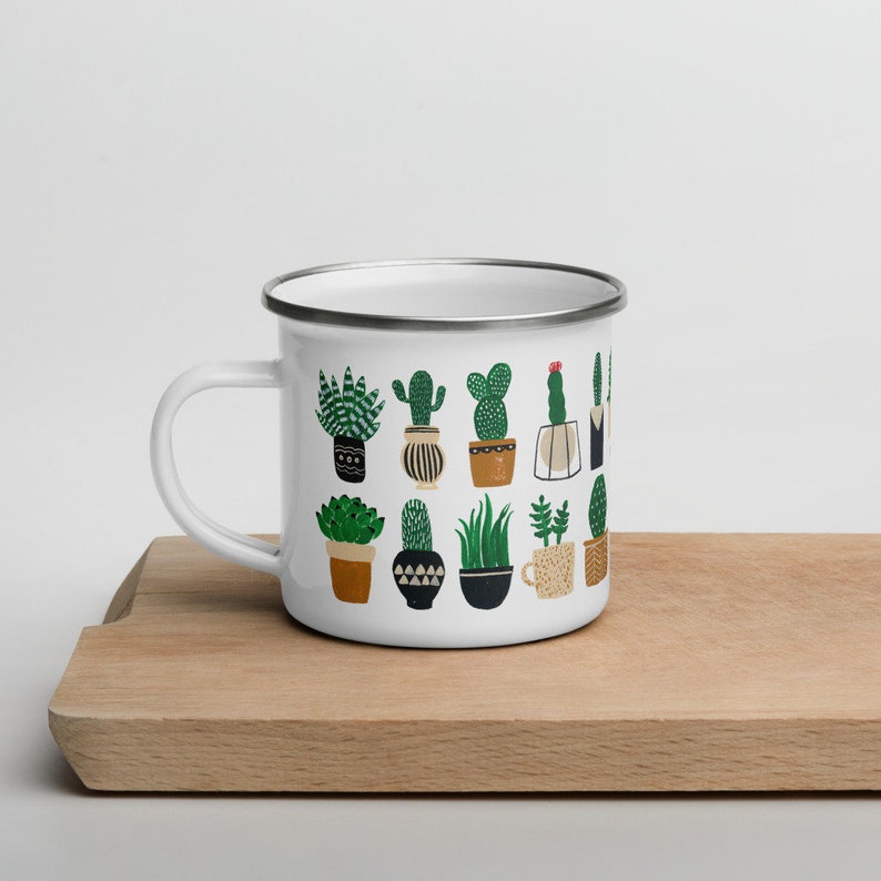 May include: White enamel mug with a colorful pattern of potted cacti and succulents. The mug has a silver rim and handle.