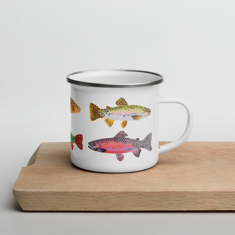 May include: A white enamel mug featuring a colorful illustration of three different fish species. The design includes a yellow-toned fish, a green-toned fish, and a pink-toned fish, all detailed with dark spots and distinct fin patterns.
