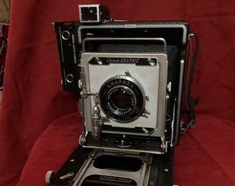 Może przedstawiać: Czarno-srebrny aparat fotograficzny Graflex Crown Graphic z wysuniętym mieszkiem i osłoną obiektywu. Aparat stoi na czerwonej powierzchni.