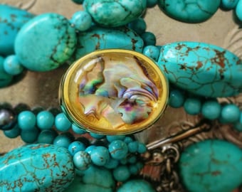 May include: A turquoise beaded necklace with a gold-rimmed oval pendant. The pendant features an iridescent shell design. The necklace includes various sizes of turquoise beads and a silver clasp.