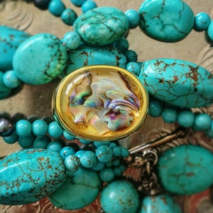 May include: A turquoise beaded necklace with a gold-rimmed oval pendant. The pendant features an iridescent shell design. The necklace includes various sizes of turquoise beads and a silver clasp.