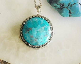 Puede incluir: Un collar con colgante plateado con una piedra turquesa redonda. La piedra tiene un color azul-verdoso moteado con inclusiones marrones. El colgante cuelga de una cadena fina. Un trozo de piedra turquesa es visible en el fondo.
