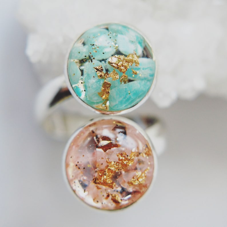 May include: Two silver rings with round, clear resin insets. The top ring has a turquoise blue resin with gold flecks. The bottom ring has a pink resin with gold flecks.