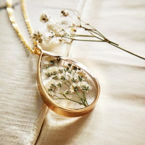 May include: A gold chain necklace with a teardrop-shaped pendant. The pendant is filled with clear resin and contains a small cluster of white dried flowers.