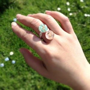 May include: Two silver rings with round resin insets. The left ring has a light green resin with gold flecks. The right ring has a light pink resin with gold flecks.