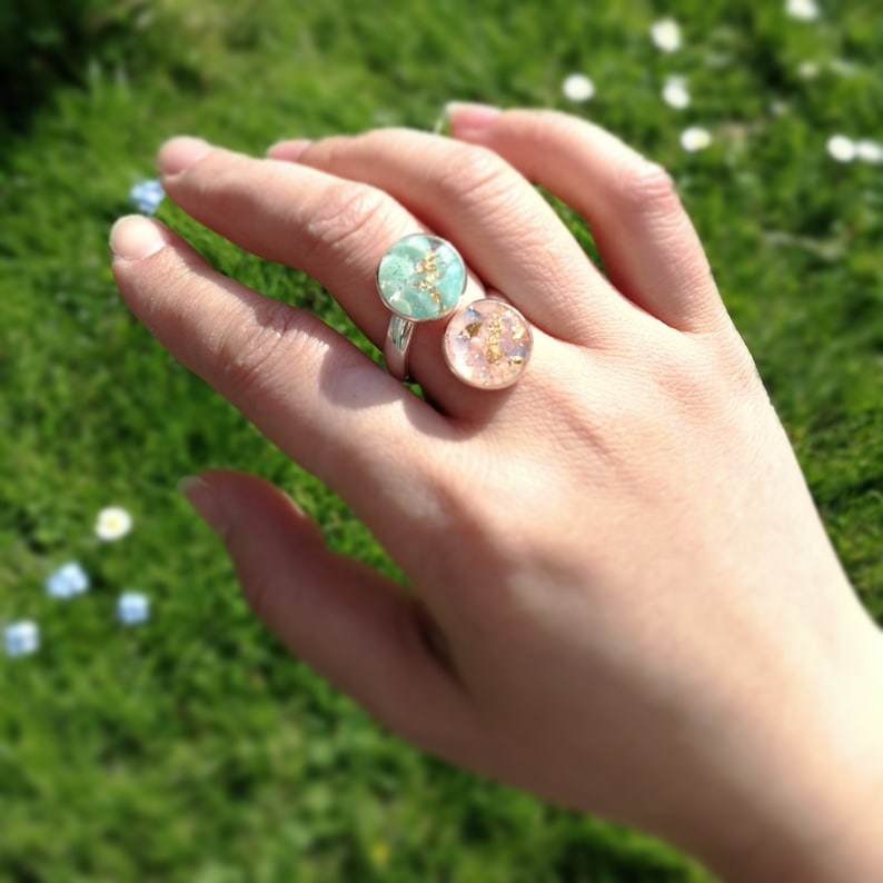 May include: Two silver rings with round resin insets. The left ring has a light green resin with gold flecks. The right ring has a light pink resin with gold flecks.