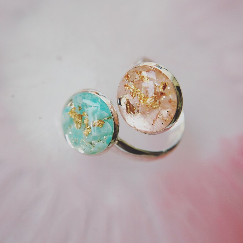 May include: A silver ring with two round resin stones. One stone is light blue with gold flecks and the other is light pink with gold flecks.