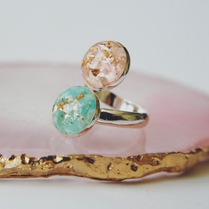 May include: A silver ring with two round resin stones, one pink and one turquoise, both with gold flecks. The ring is on a pink and gold agate coaster.