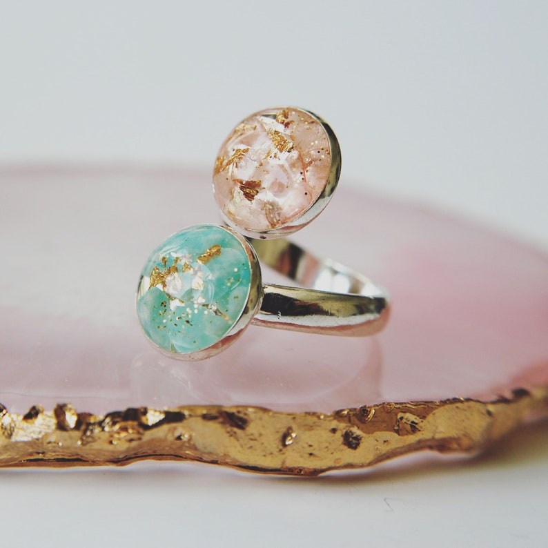 May include: A silver ring with two round resin stones, one pink and one turquoise, both with gold flecks. The ring is on a pink and gold agate coaster.