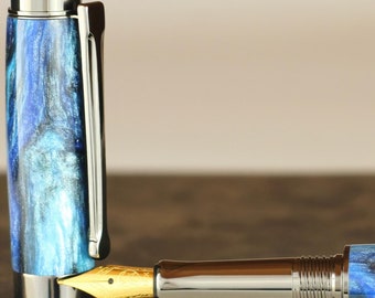 Könnte beinhalten: Ein blauer und schwarzer Muster-Füllfederhalter mit einer goldenen Feder und einer silbernen Kappe. Der Stift liegt auf einem braunen Leder-Notizbuch.
