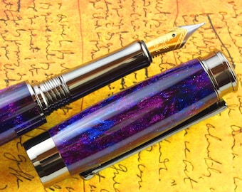May include: A purple and blue fountain pen with a gold-toned nib and a silver-toned body. The pen is lying on a piece of paper with handwritten text.