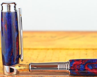 Könnte beinhalten: Ein Füllfederhalter mit einem blauen und roten Marmormuster und einer goldfarbenen Feder. Der Stift ist geschlossen und liegt auf einem Buch.