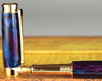 Könnte beinhalten: Ein violetter und blauer Füllfederhalter mit goldenen Akzenten. Der Stift ist aus Holz und hat ein einzigartiges Marmormuster.