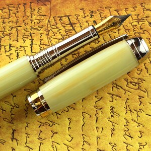May include: A gold and cream colored fountain pen with a gold trim. The pen is lying on a piece of paper with writing on it.