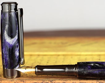 Könnte beinhalten: Ein luxuriöser Füllfederhalter mit einem schwarz-violett marmorierten Design. Der Stift hat eine goldene Feder und einen silbernen Clip. Der Stift liegt auf einem Vintage-Buch.