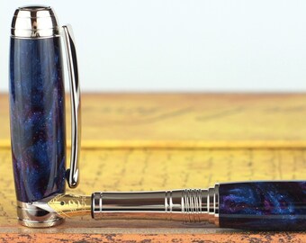 Könnte beinhalten: Ein blauer und violetter Füllfederhalter mit silbernen Akzenten und einem wirbelnden Muster. Der Stift liegt auf einem braunen Buch.