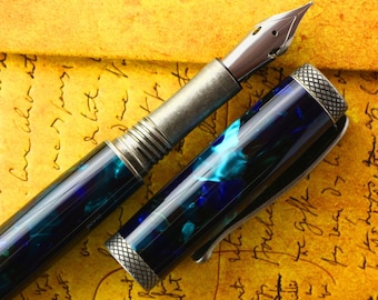 Könnte beinhalten: Ein blauer und grüner Füllfederhalter mit einem silbernen Metallkörper und einer silbernen Metallkappe. Der Stift liegt auf einem Blatt Papier mit handschriftlichem Text.